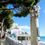 An ancient stone column on the Amalfi coast under a bright blue sky, framed by green Italian stone pines (pinie).