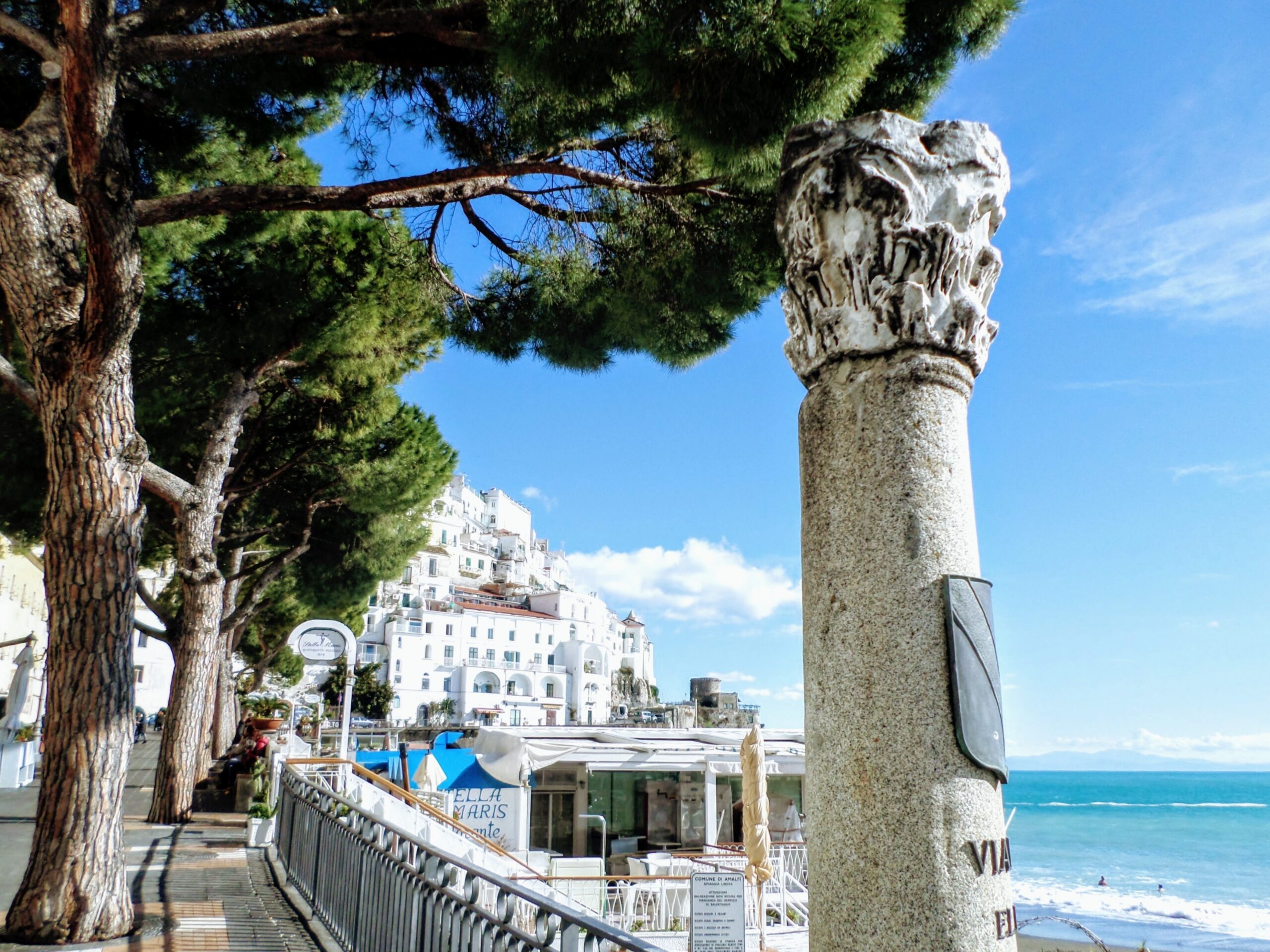 An ancient stone column on the Amalfi coast under a bright blue sky, framed by green Italian stone pines (pinie).