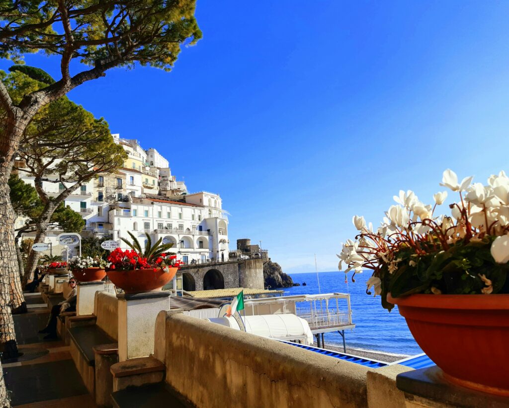 A vibrant summer promenade in Amalfi filled with blooming flowers, sun-drenched coastal architecture, and the blue Mediterranean Sea.